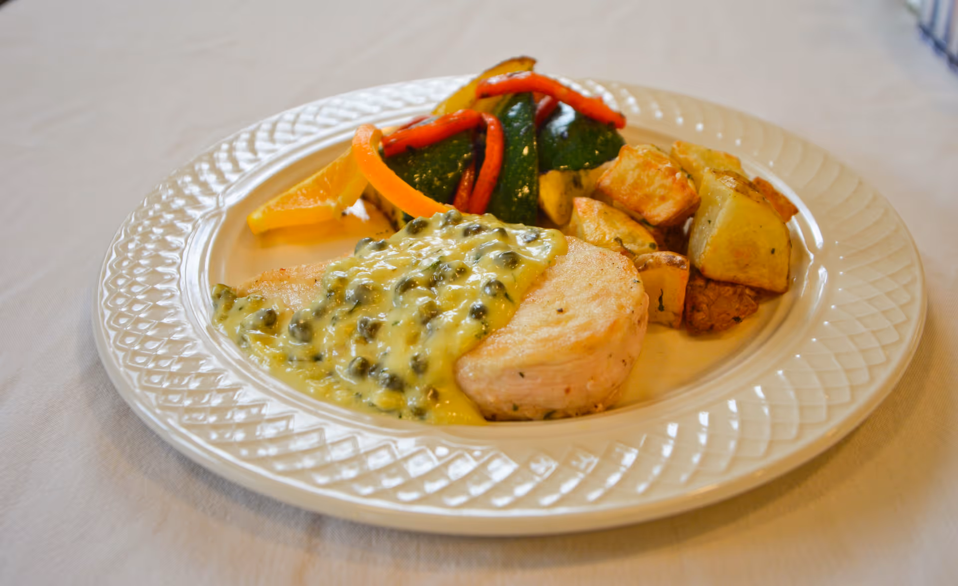 A white plate with a serving of cooked chicken breast topped with a creamy caper sauce, accompanied by roasted potatoes and sautéed vegetables including zucchini and red bell peppers, placed on a white tablecloth.