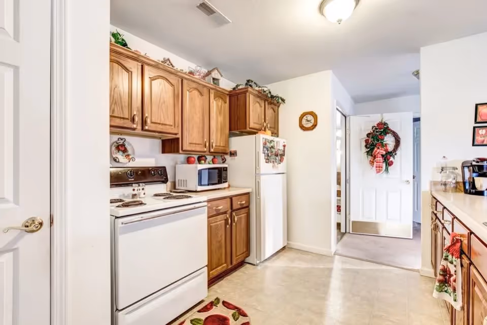 A bright kitchen with wooden cabinets, a white stove, microwave, and refrigerator. The kitchen has a tiled floor and a door decorated with a festive wreath. There are small decorative items on top of the cabinets and a clock on the wall.