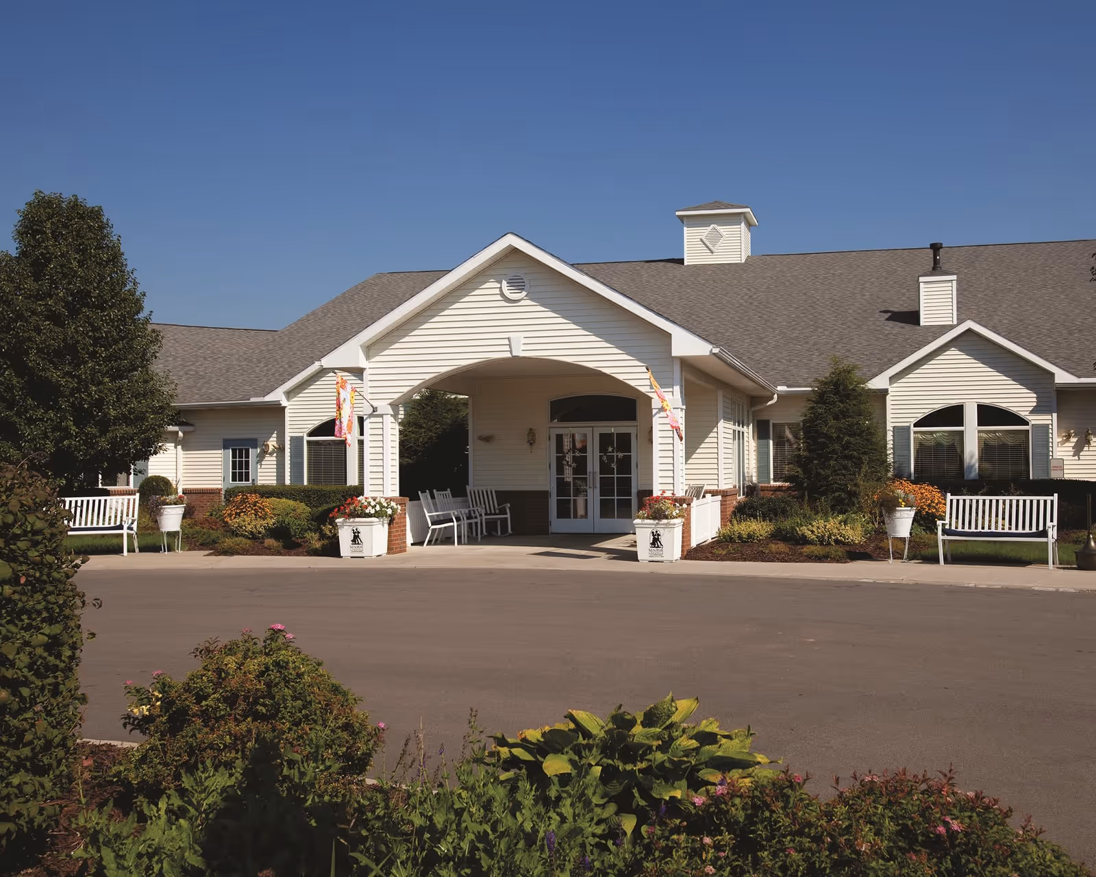 Exterior view of a single-story building with white siding and a gray roof under a clear blue sky. The entrance features a covered porch with white columns, benches, and flower pots on either side. There are bushes, trees, and landscaped plants around the building.