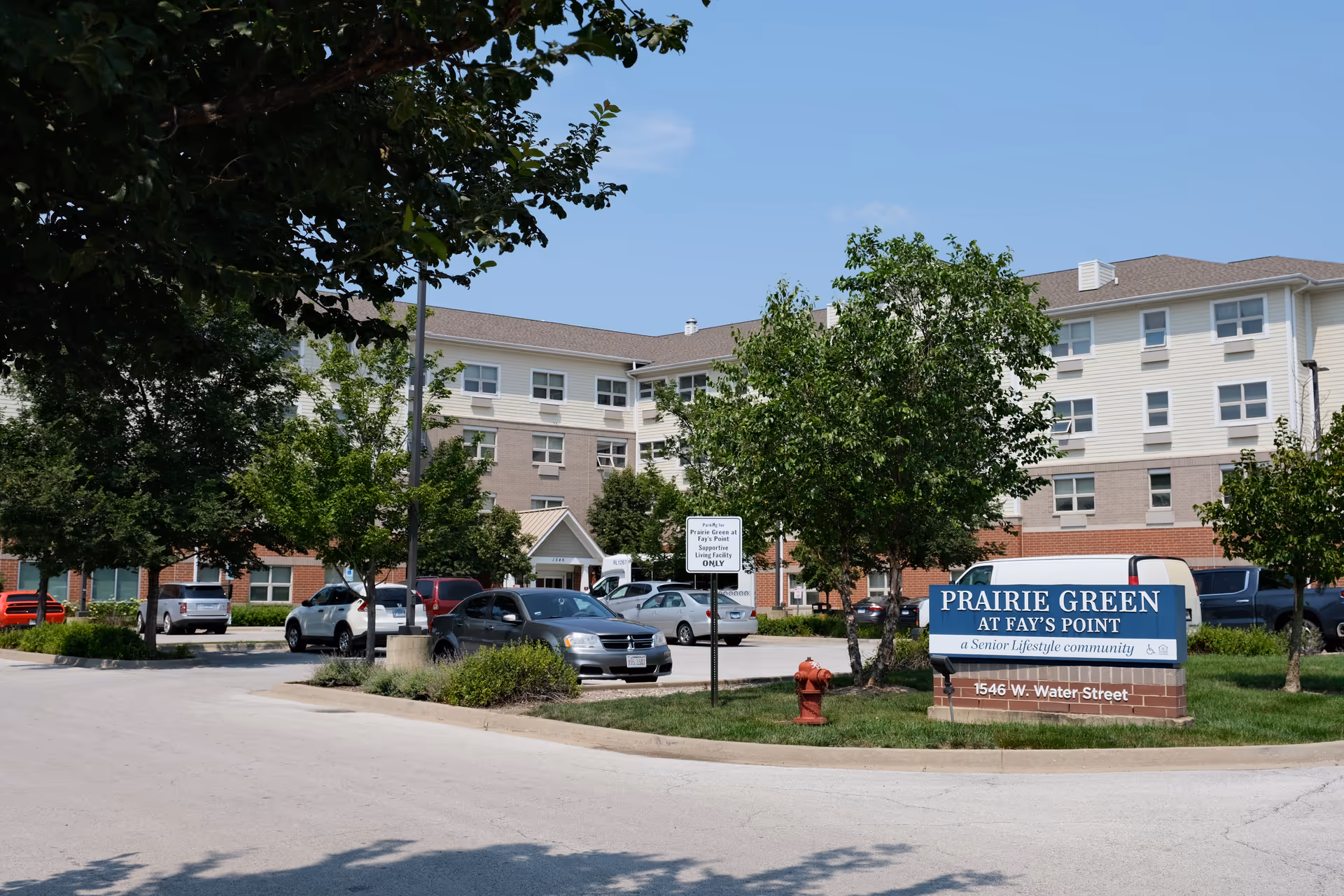 Exterior view of Prairie Green at Fay's Point senior living community building with a parking lot in front, several cars parked, trees, and a blue sign displaying the community name and address 1546 W. Water Street.