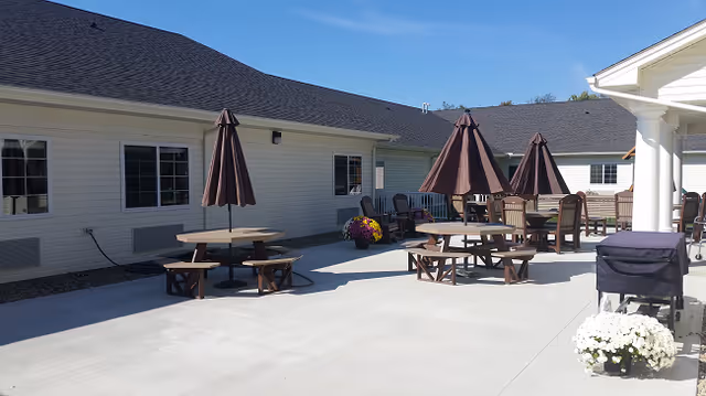 Outdoor patio area at Centreville Village Assisted Living with several round picnic tables, each shaded by a closed brown umbrella. There are additional chairs and a barbecue grill on the concrete patio, adjacent to a light-colored building under a clear blue sky.