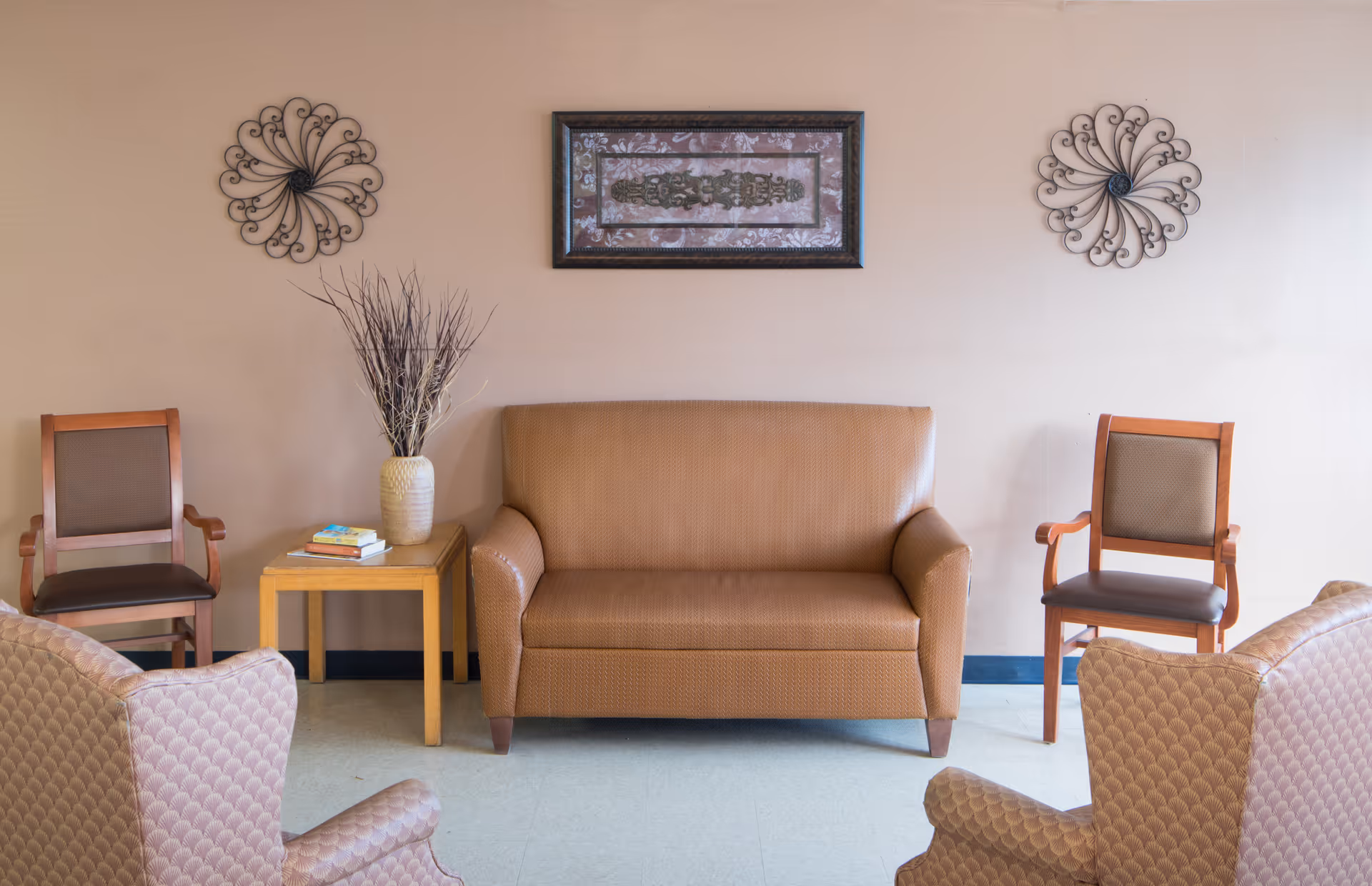 A cozy seating area with a brown loveseat centered against a beige wall, flanked by two wooden chairs with cushioned seats. A small wooden side table with a vase of dried branches and a few books is placed to the left of the loveseat. Two decorative metal wall hangings and a framed artwork are mounted on the wall above the loveseat.