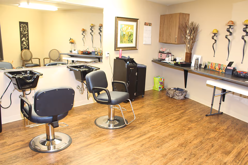 Interior of a senior living community salon area with two black salon chairs in front of two black hair washing sinks. A large mirror is mounted on the wall behind the chairs. The room has wooden flooring, a countertop with various items, a wall cabinet, and decorative wall art.