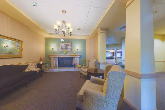 A cozy living room area in an assisted living facility featuring a fireplace with a painting above it, two armchairs and a round wooden table, a dark sofa with a blanket, a large mirror on the wall, and soft lighting from a chandelier and wall sconces.