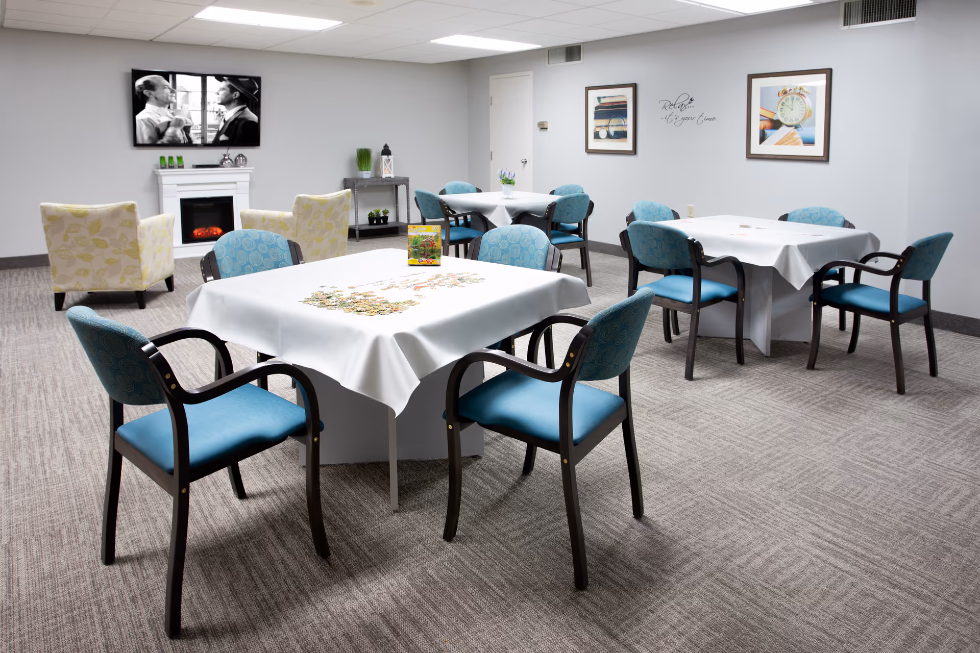 A bright communal dining/activity room with several tables covered in white tablecloths, blue chairs, a TV above a fireplace and armchairs.