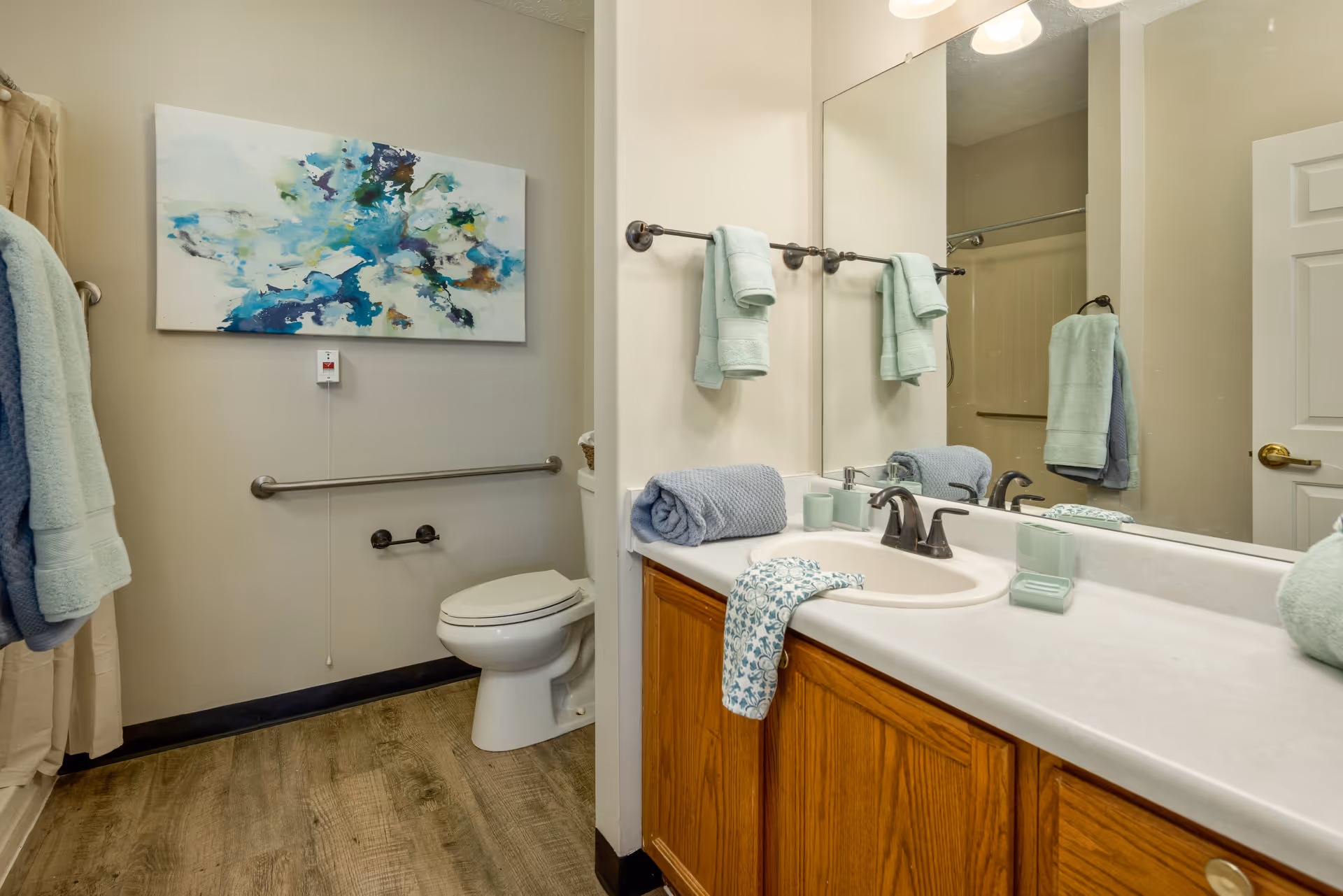 A clean bathroom with a white toilet, a wooden vanity with a white countertop and sink, a large mirror above the sink, and several light blue towels hanging on towel racks and placed on the countertop. There is a colorful abstract painting on the wall above the toilet and a shower curtain partially visible on the left side.