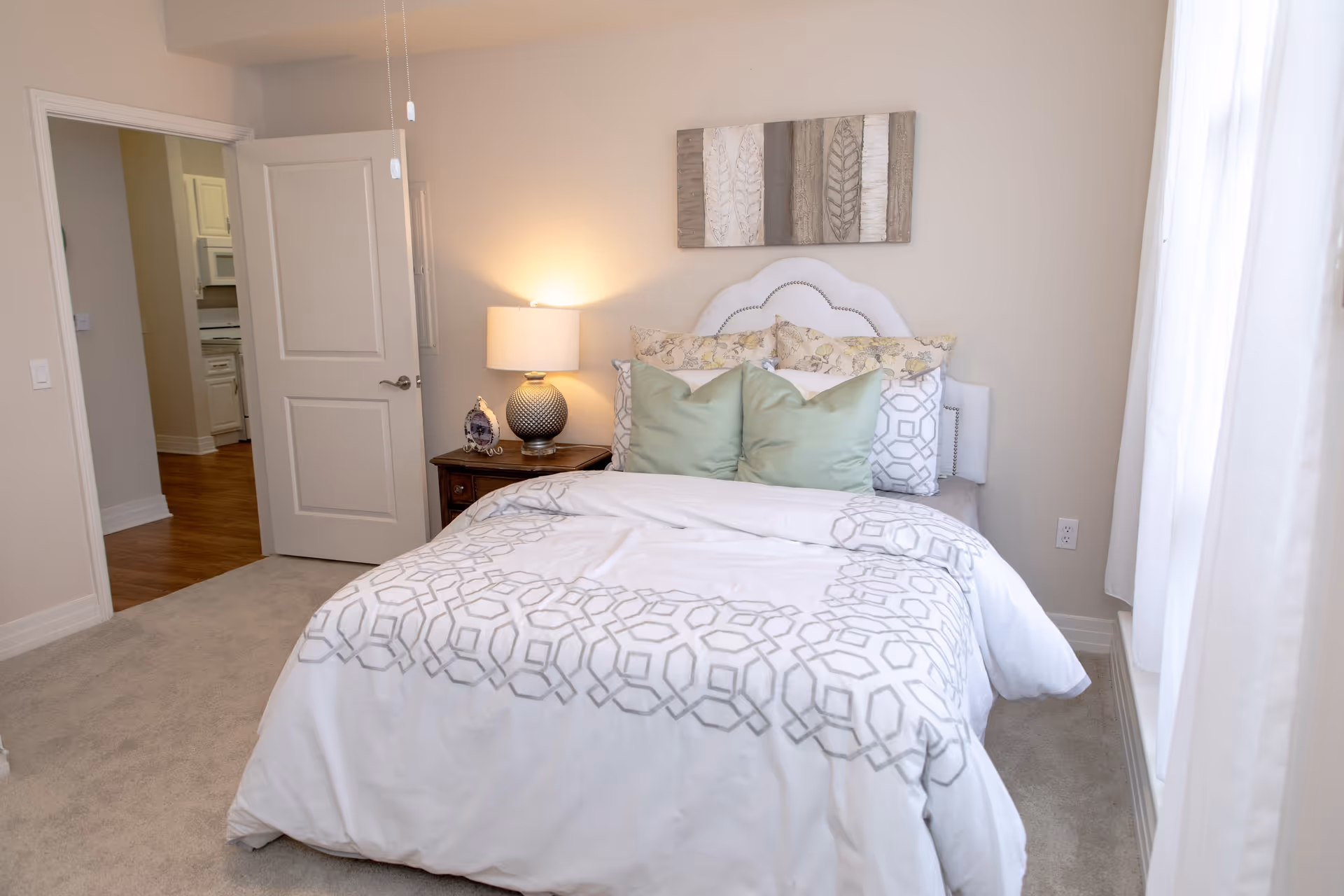 A cozy bedroom with a neatly made bed featuring white and gray patterned bedding and multiple pillows. There is a wooden nightstand with a decorative lamp and a small clock. The room has beige walls, a piece of artwork above the bed, and a window with white curtains letting in natural light. An open door reveals a glimpse of a kitchen area with wooden flooring.