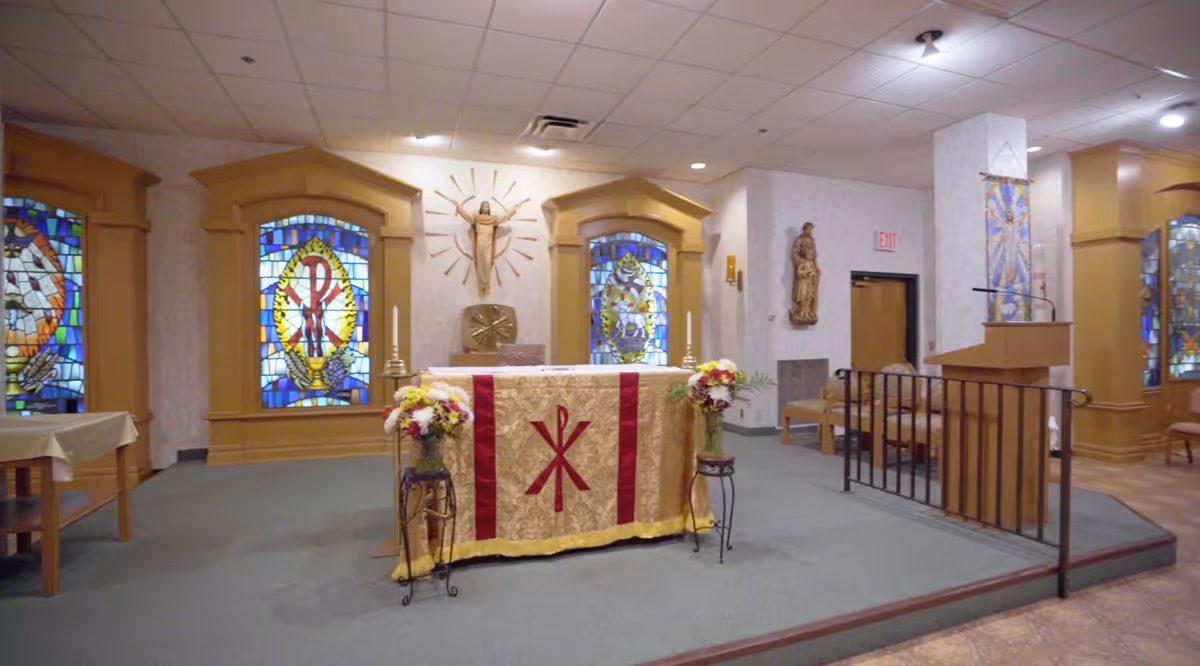 Interior view of a chapel with an altar covered in a gold and red cloth featuring a Christian Chi Rho symbol. The altar is flanked by flower arrangements on stands. Behind the altar are three stained glass windows with religious symbols and a crucifix mounted on the wall. There is a lectern to the right and wooden chairs along the walls.