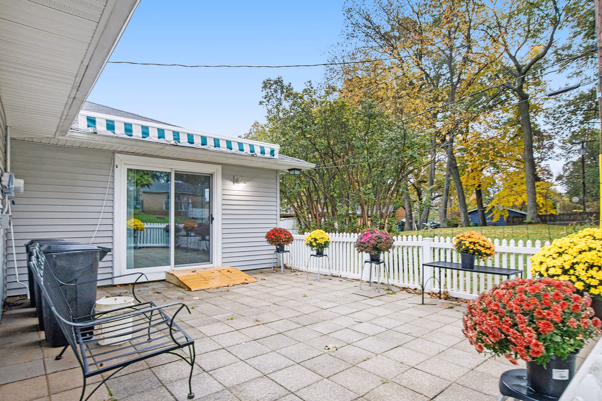 Outdoor patio area with tiled flooring, a black metal bench, several potted colorful flowers on stands and tables, a white picket fence, and a sliding glass door leading into the building. Trees with autumn foliage are visible in the background under a clear blue sky.