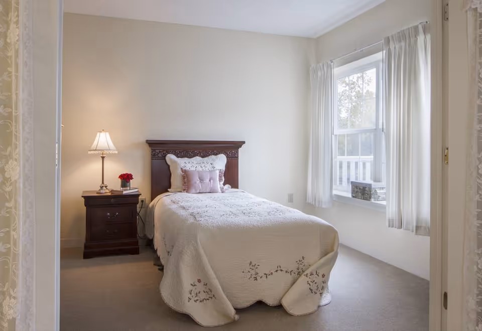 A cozy bedroom with a single bed covered in a white quilt with floral embroidery. The bed has a wooden headboard and is adorned with decorative pillows. To the left of the bed is a dark wooden nightstand with a table lamp and a small vase of red flowers. A window with white curtains allows natural light to brighten the room.