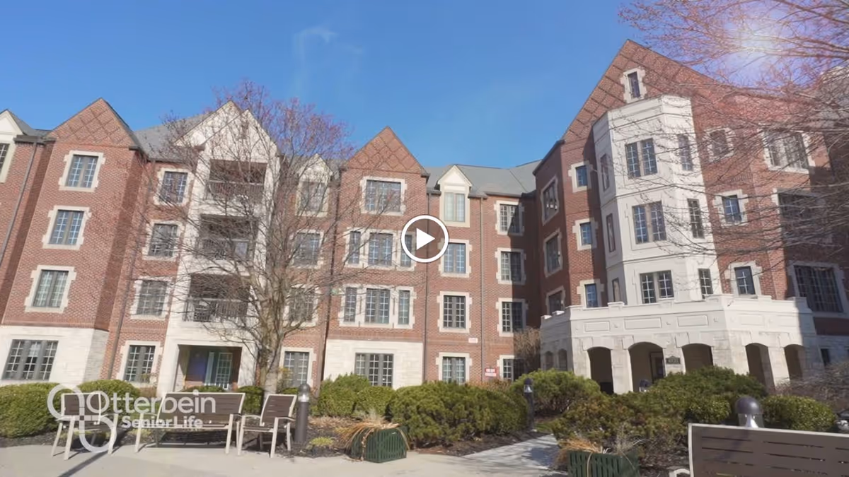 Exterior view of a multi-story senior living community building with brick and stone facade, multiple windows, and a small outdoor seating area with chairs and a table surrounded by bushes and trees under a clear blue sky.