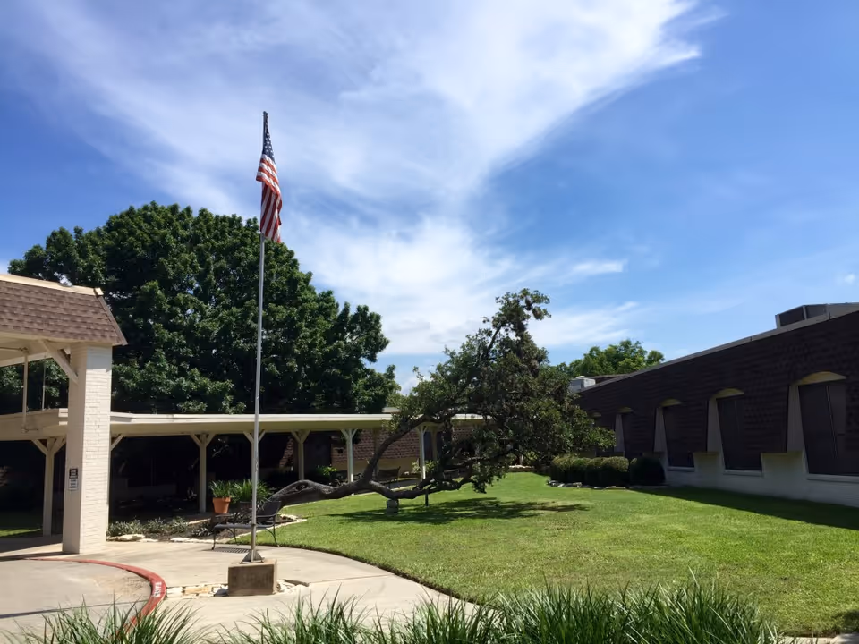 Courtyard and entrance of a senior living facility with an American flag, a leaning tree, covered walkway and lawn under a blue sky.