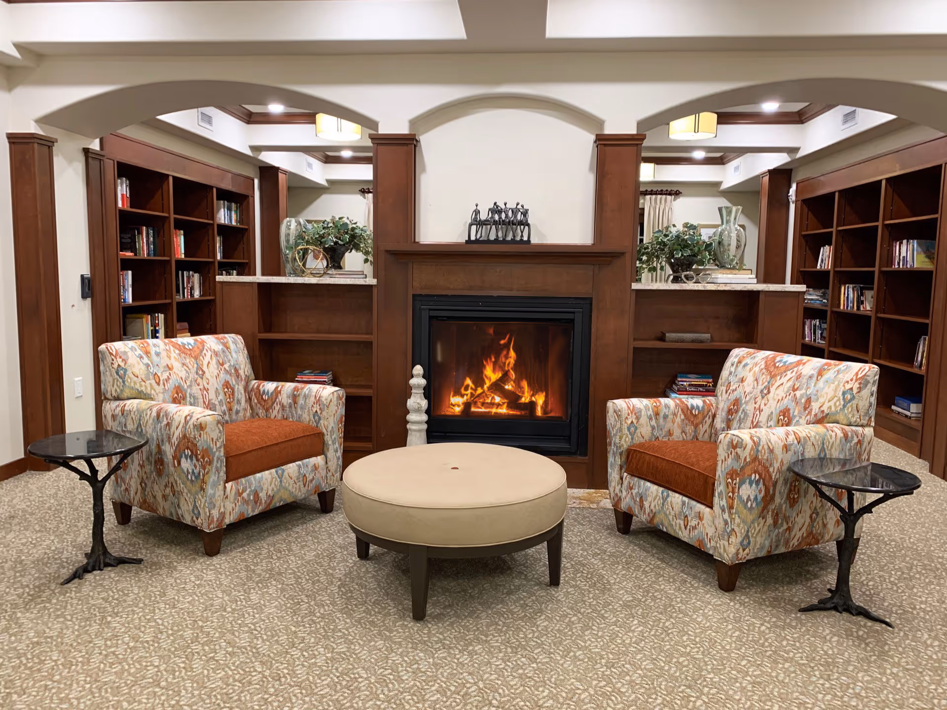 A cozy living room area with two patterned armchairs featuring orange cushions, positioned on either side of a round beige ottoman. Behind the ottoman is a wooden fireplace with a fire burning inside. The fireplace is flanked by built-in wooden bookshelves filled with books and decorative items. The room has a beige carpet and light-colored walls with wooden trim and arches.