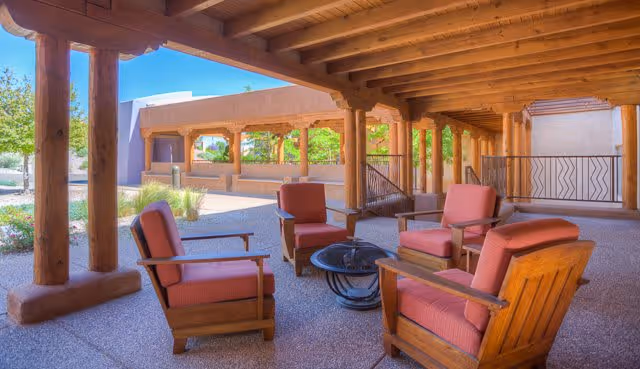 Outdoor covered patio area with wooden beams and columns, featuring four cushioned wooden armchairs arranged around a circular fire pit table. The background shows a landscaped garden area with trees and shrubs under a clear blue sky.