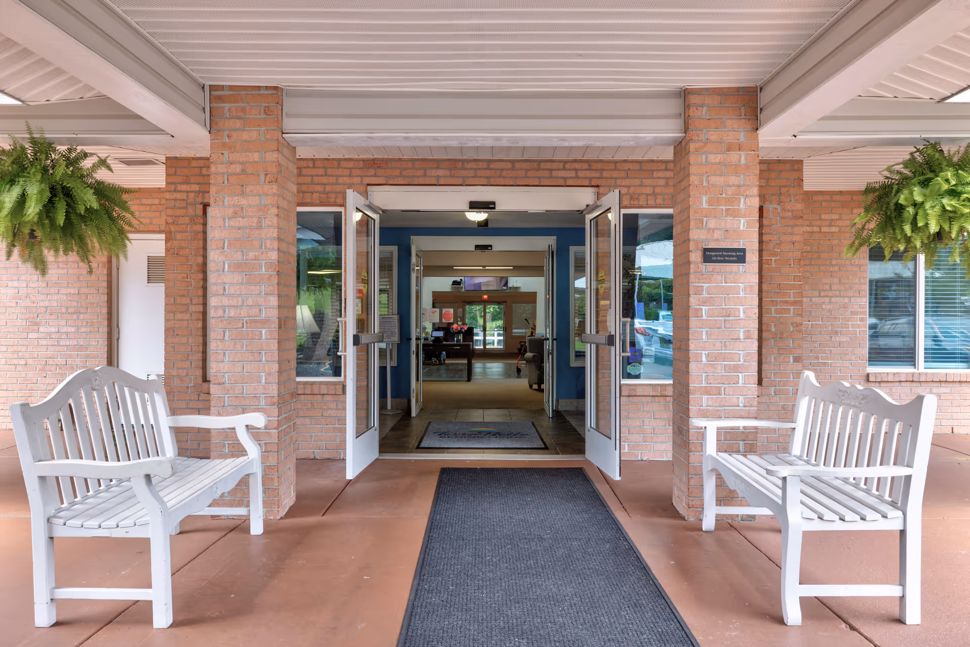 Entrance to a building with double glass doors open, flanked by two white wooden benches and hanging green ferns on either side. The building has brick walls and a covered walkway with a black mat leading to the doors.