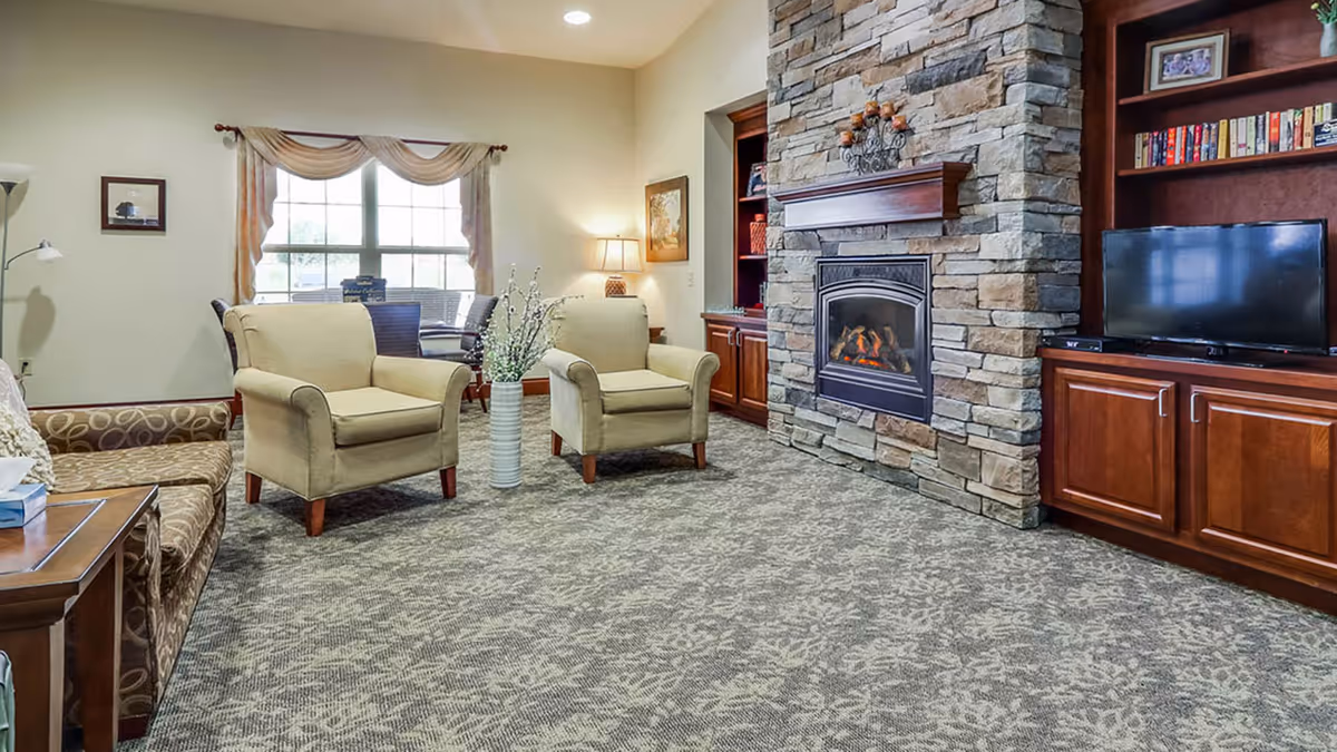 A cozy living room with two beige armchairs and a patterned sofa arranged around a stone fireplace with a lit fire. There is a TV on a wooden cabinet to the right, a window with draped curtains in the background, and a small dining table with chairs near the window. The room has carpeted flooring and warm lighting from a table lamp.