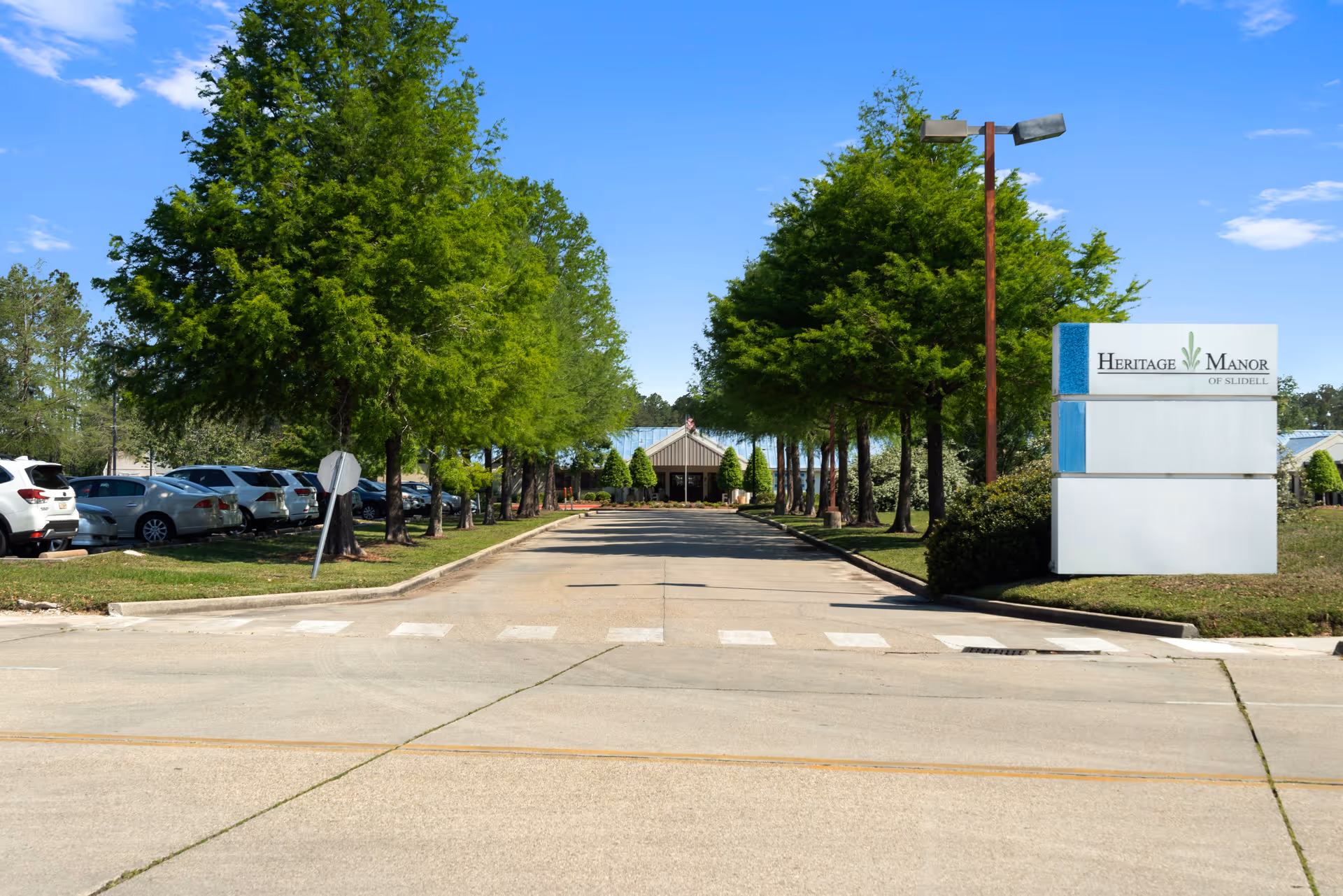 Driveway lined with trees leading to the entrance of Heritage Manor Slidell, with a facility sign on the right and parked cars on the left.