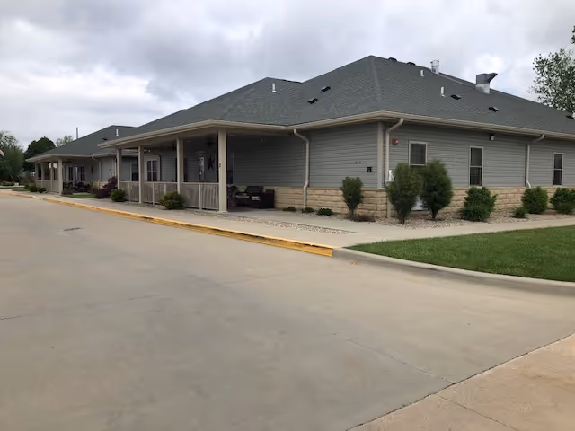 Exterior view of a single-story building with gray siding and a dark gray roof, featuring a covered porch area with seating. The building is surrounded by a concrete driveway and small landscaped areas with bushes and grass under a cloudy sky.