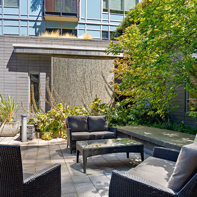 Outdoor courtyard with wicker seating, a glass-top coffee table, lush plants and a textured water feature set against a modern building facade.