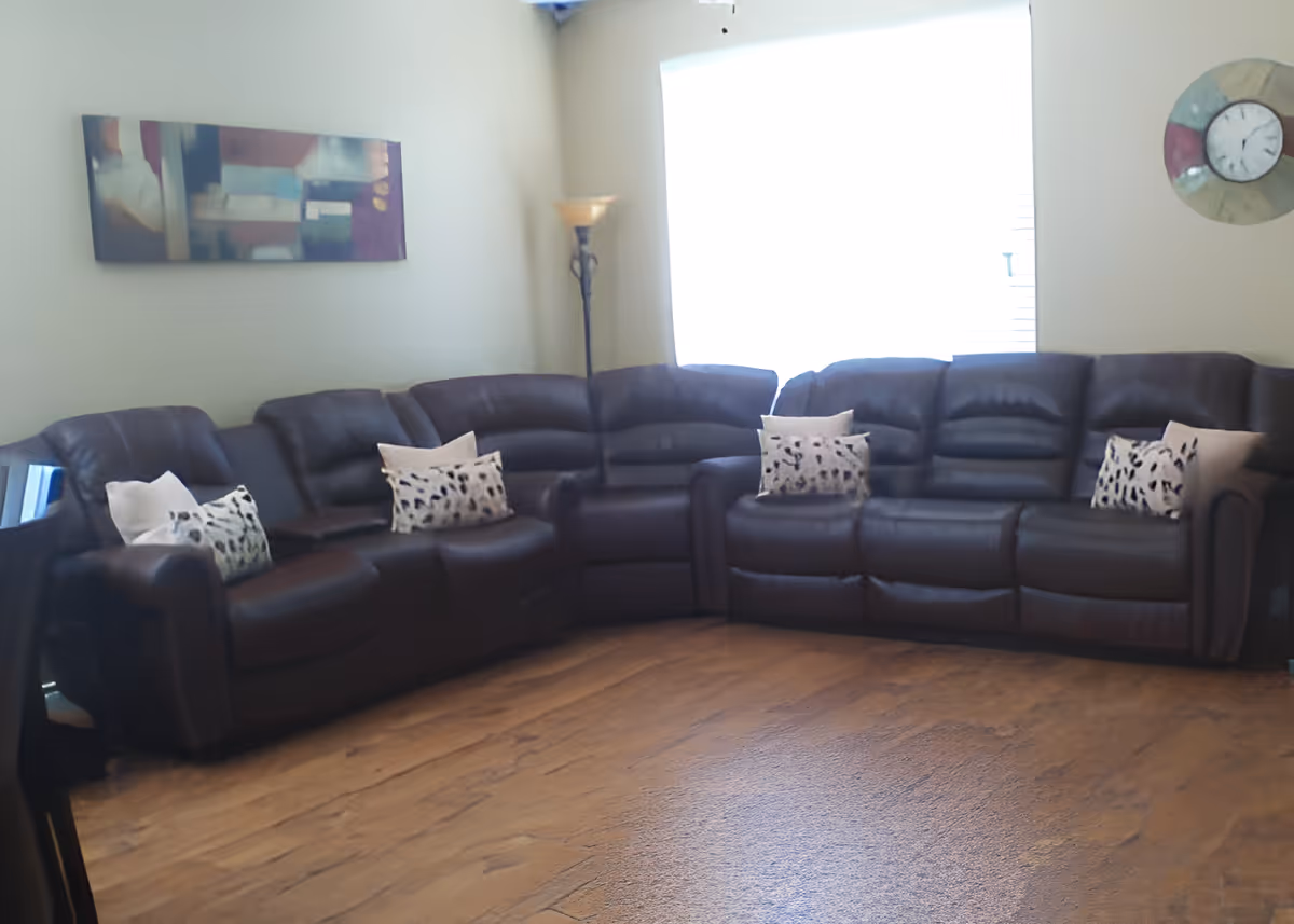 A living room with two dark brown leather sofas arranged in an L-shape, each sofa adorned with patterned throw pillows. There is a tall floor lamp in the corner and a large window with blinds letting in bright light. The walls are light-colored and decorated with a rectangular abstract painting and a round clock.
