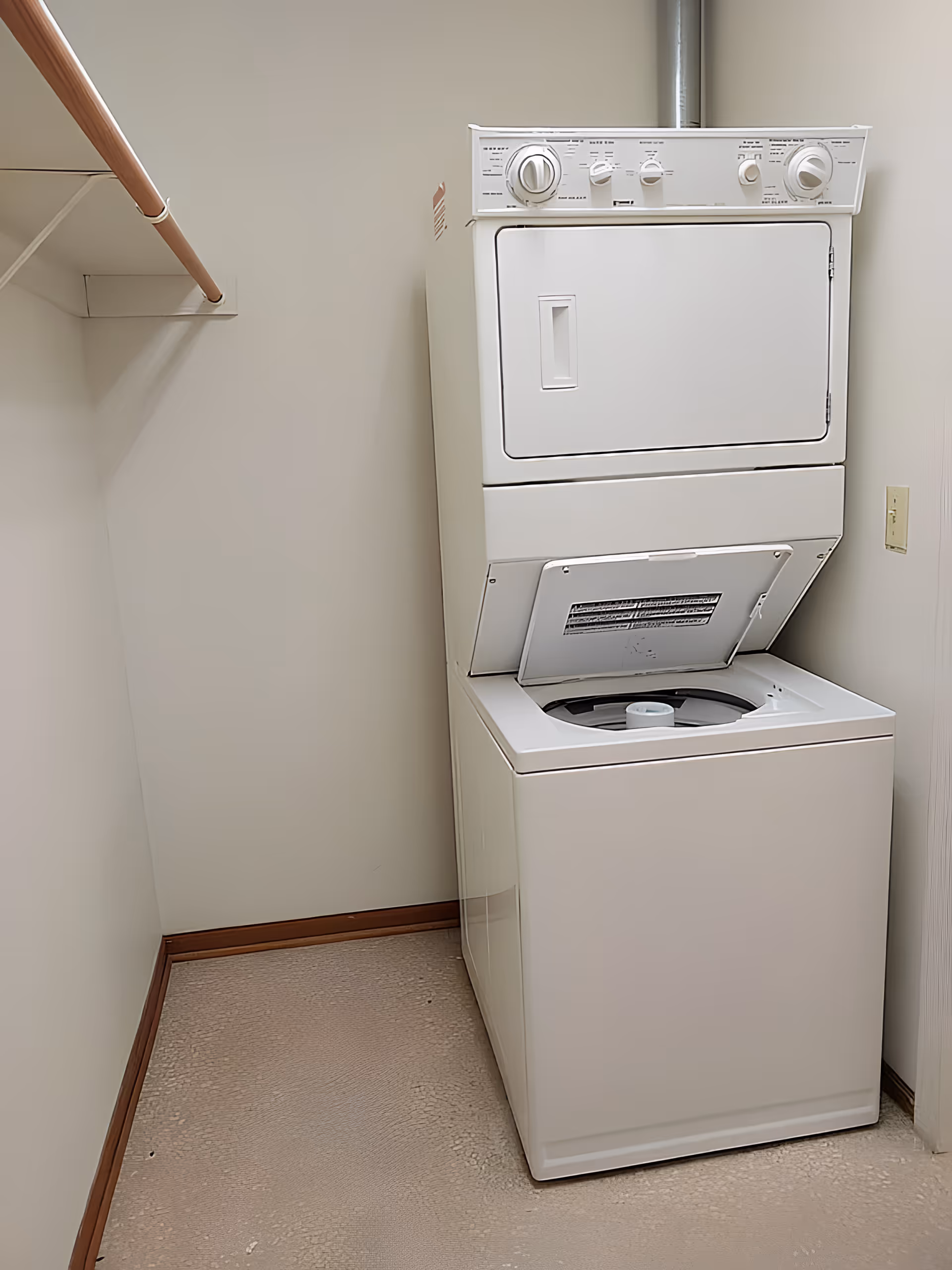 A small laundry room with a stacked white washer and dryer unit. The washer lid is open, and there is a wooden hanging rod on the left wall. The walls are plain and light-colored, and the floor has a textured surface.