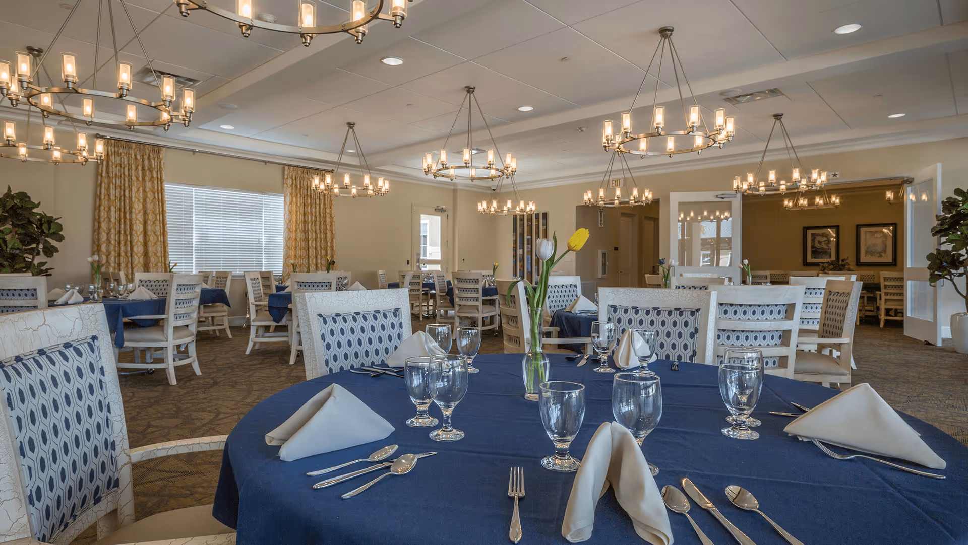 A spacious dining room with multiple round tables covered in blue tablecloths, each set with folded white napkins, glassware, and silverware. The chairs have patterned upholstery, and the room is well-lit with several chandeliers hanging from the ceiling. Large windows with yellow patterned curtains and potted plants add to the decor.