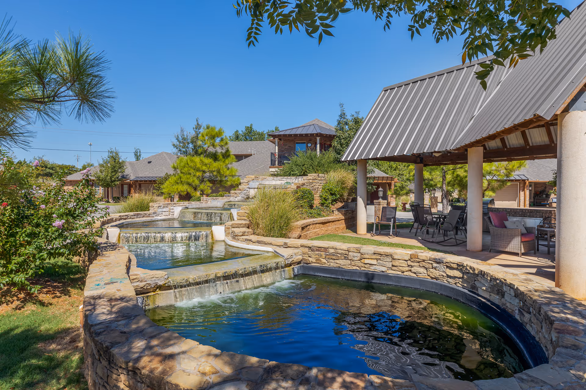 Outdoor area of a senior living facility featuring a multi-tiered stone water feature with cascading waterfalls, surrounded by greenery and trees. There is a covered patio area with chairs and tables for seating and relaxation under a metal roof. Residential buildings are visible in the background under a clear blue sky.
