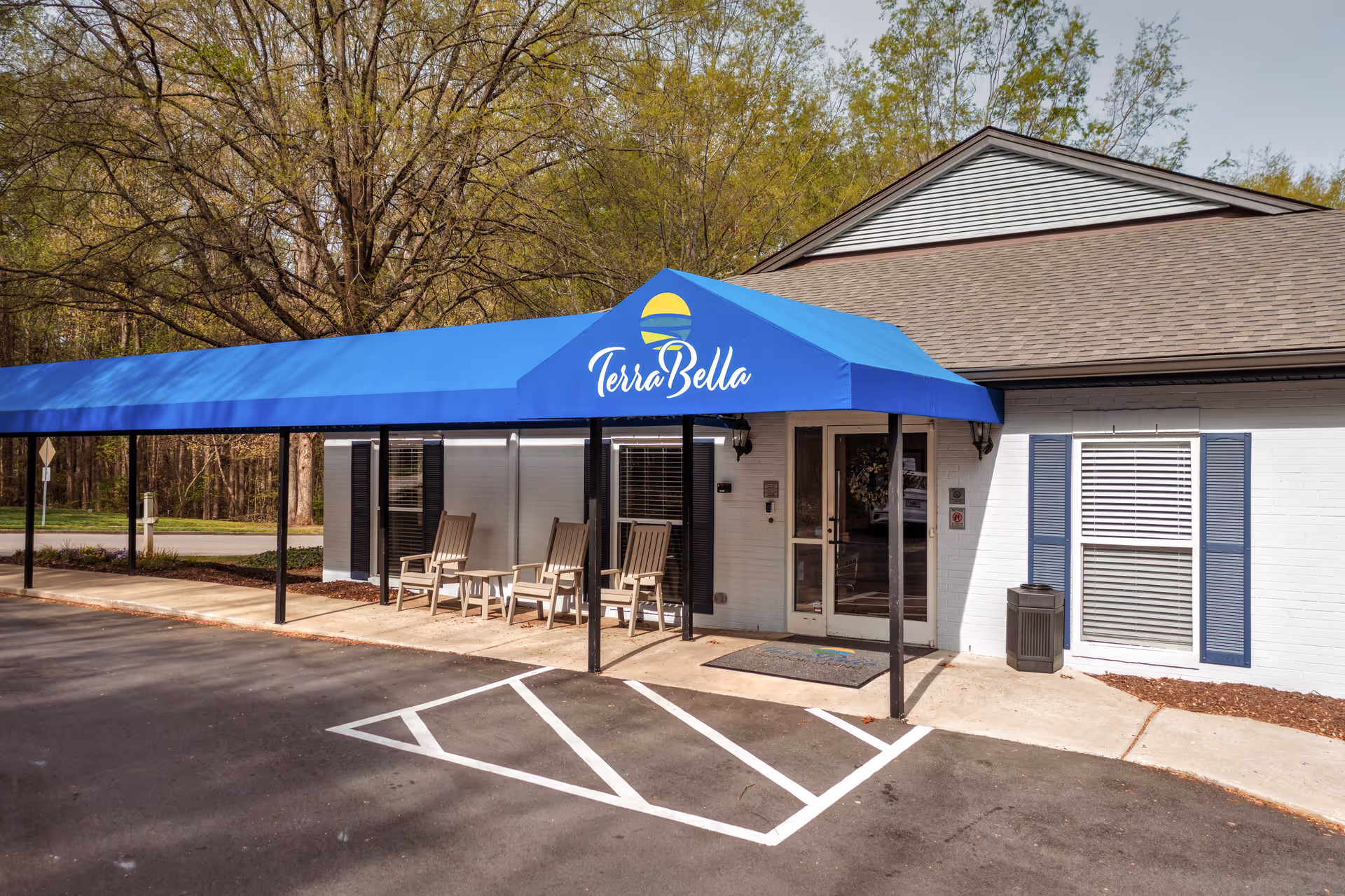 Exterior view of TerraBella Northridge facility entrance with a blue awning displaying the TerraBella logo. There are four wooden chairs on the porch under the awning, a glass door entrance, and a parking area with marked spaces in front. Trees and greenery are visible in the background.
