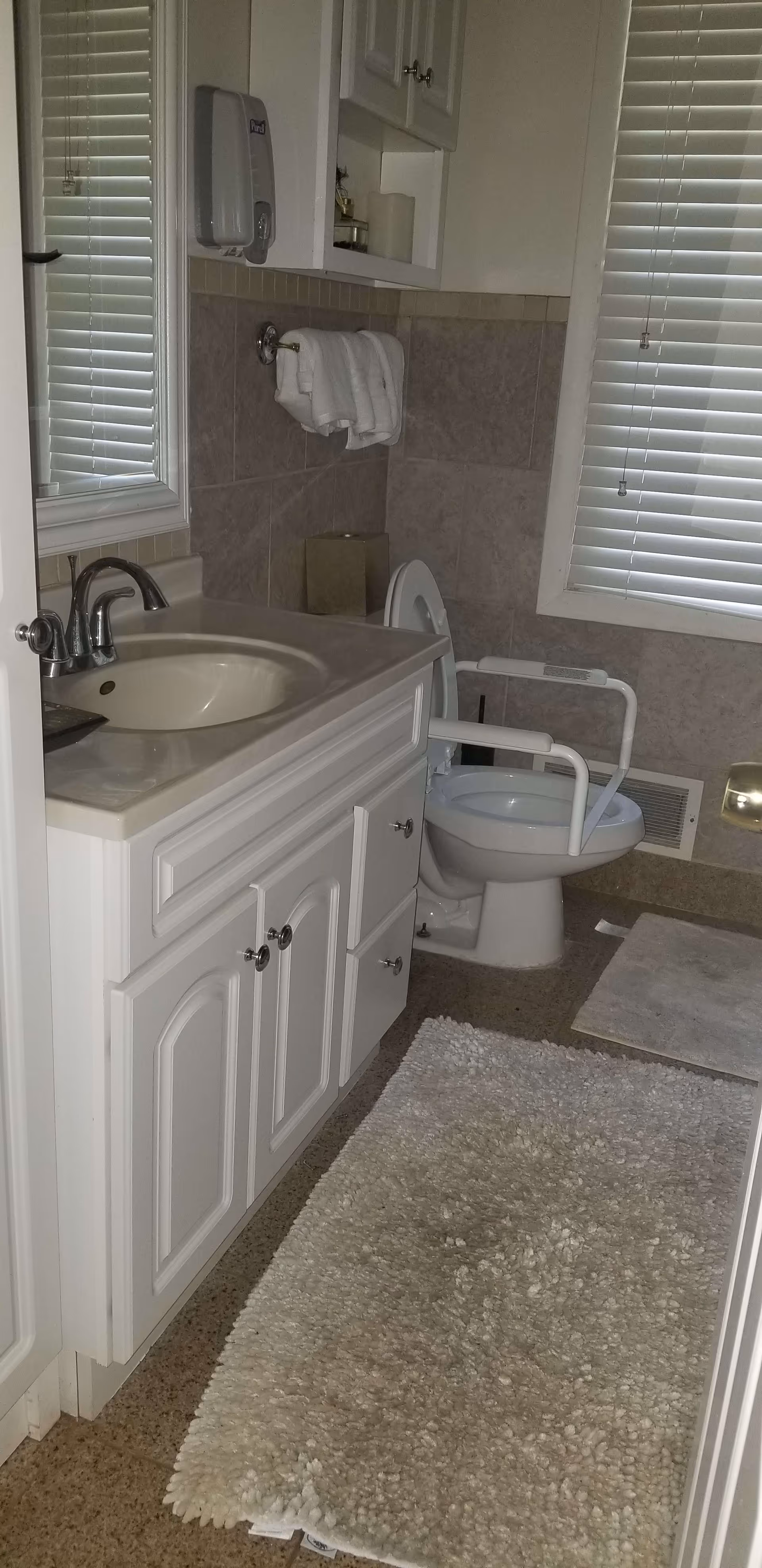 A bathroom with a white vanity and sink on the left, a mirror above the sink, a towel rack with folded white towels, a toilet with white support rails, a window with white blinds, and two white bath mats on the floor.