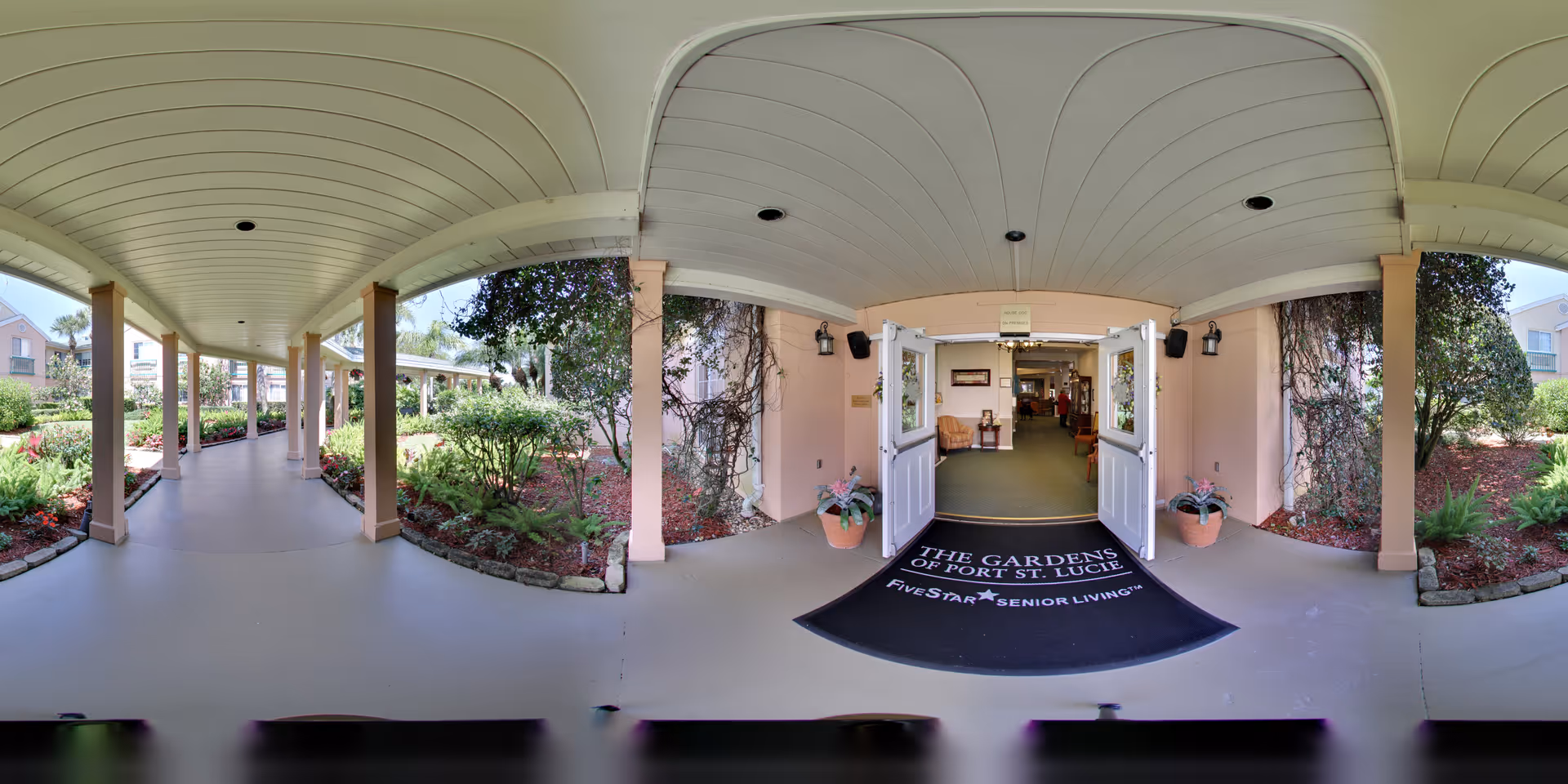 Covered walkway leading to the entrance of The Gardens of Port St. Lucie senior living facility, with open double doors and a welcome mat displaying the facility's name. The walkway is flanked by landscaped garden beds with shrubs and flowers, and the building exterior is light pink with white trim.