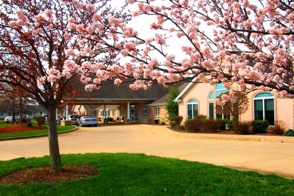 Exterior view of Brookstone Estates Of Effingham facility with a driveway, green lawn, and blooming pink cherry blossom trees in the foreground. The building has large windows and a covered entrance with a car parked nearby.