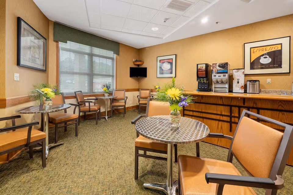 Small communal seating area with round metal-top tables, upholstered chairs, a coffee station and framed posters on the walls.