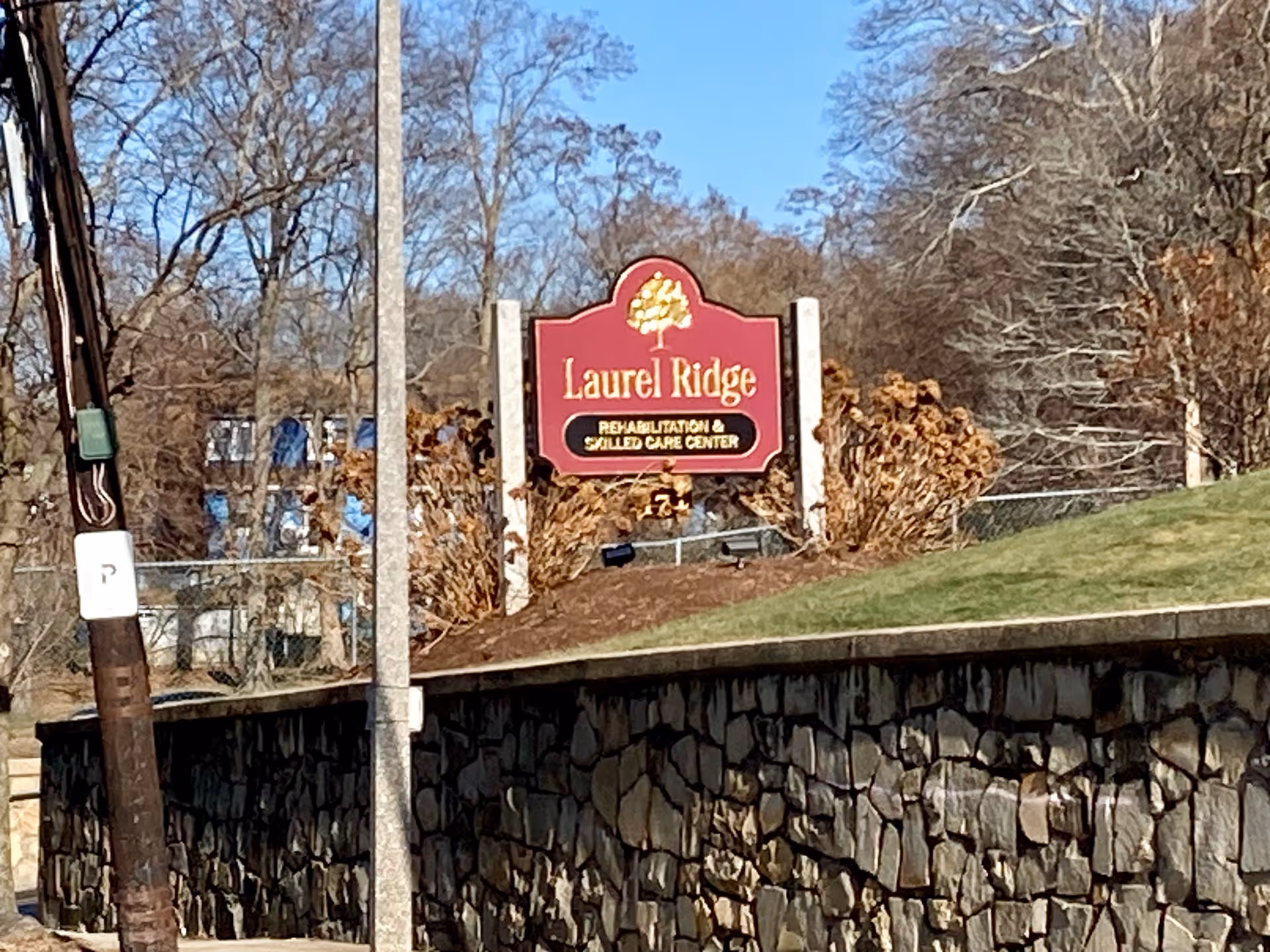 Red Laurel Ridge Rehabilitation & Skilled Care Center sign on a grassy hill above a stone retaining wall with trees in the background.