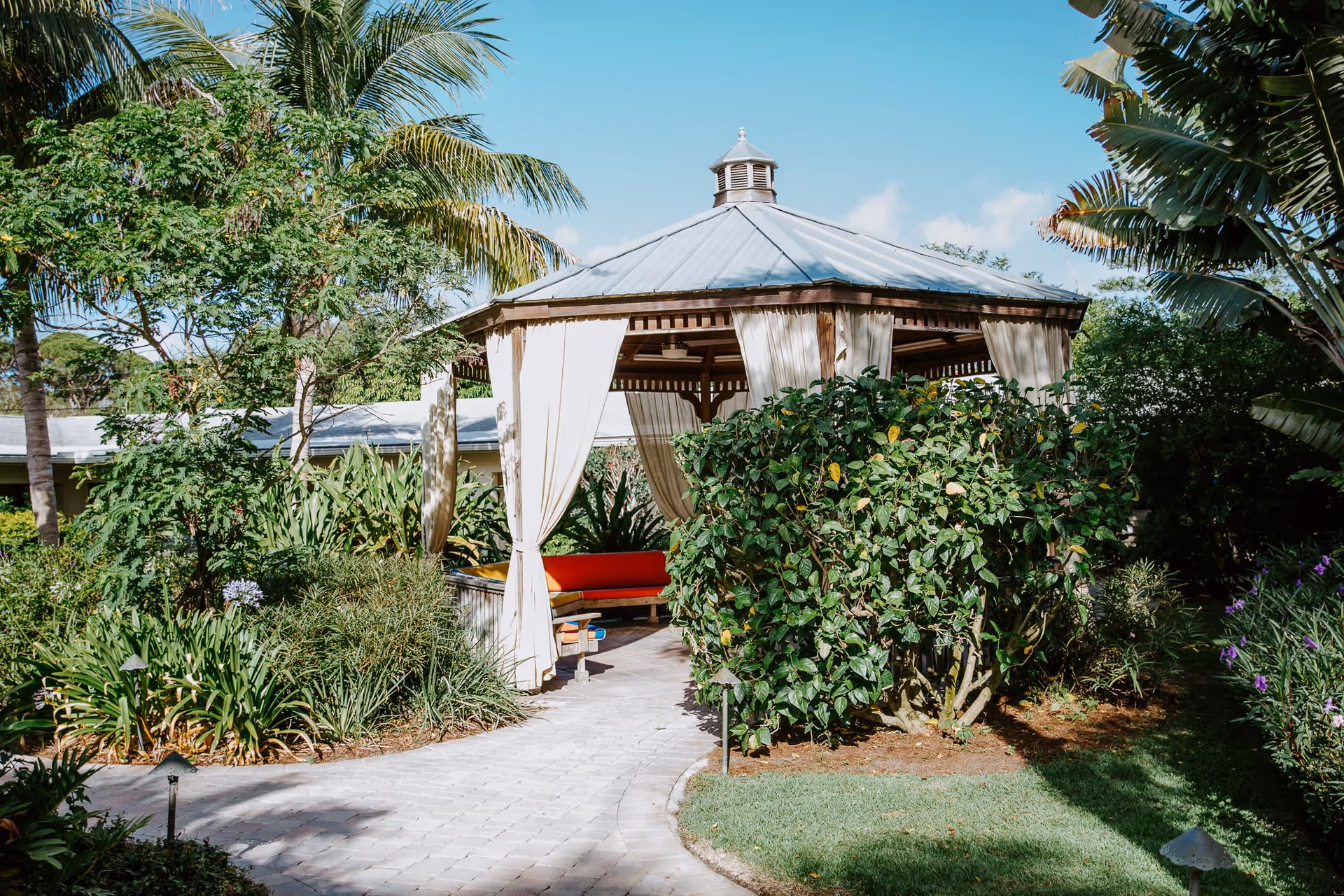 A gazebo with a metal roof and white curtains tied back, surrounded by lush green plants and palm trees, with a paved walkway leading to it under a clear blue sky.