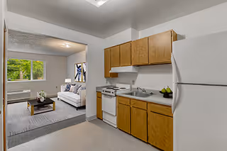 View of a small kitchen area with wooden cabinets, a white refrigerator, stove, and sink. The kitchen opens into a living room with a white sofa, coffee table, and a window letting in natural light.