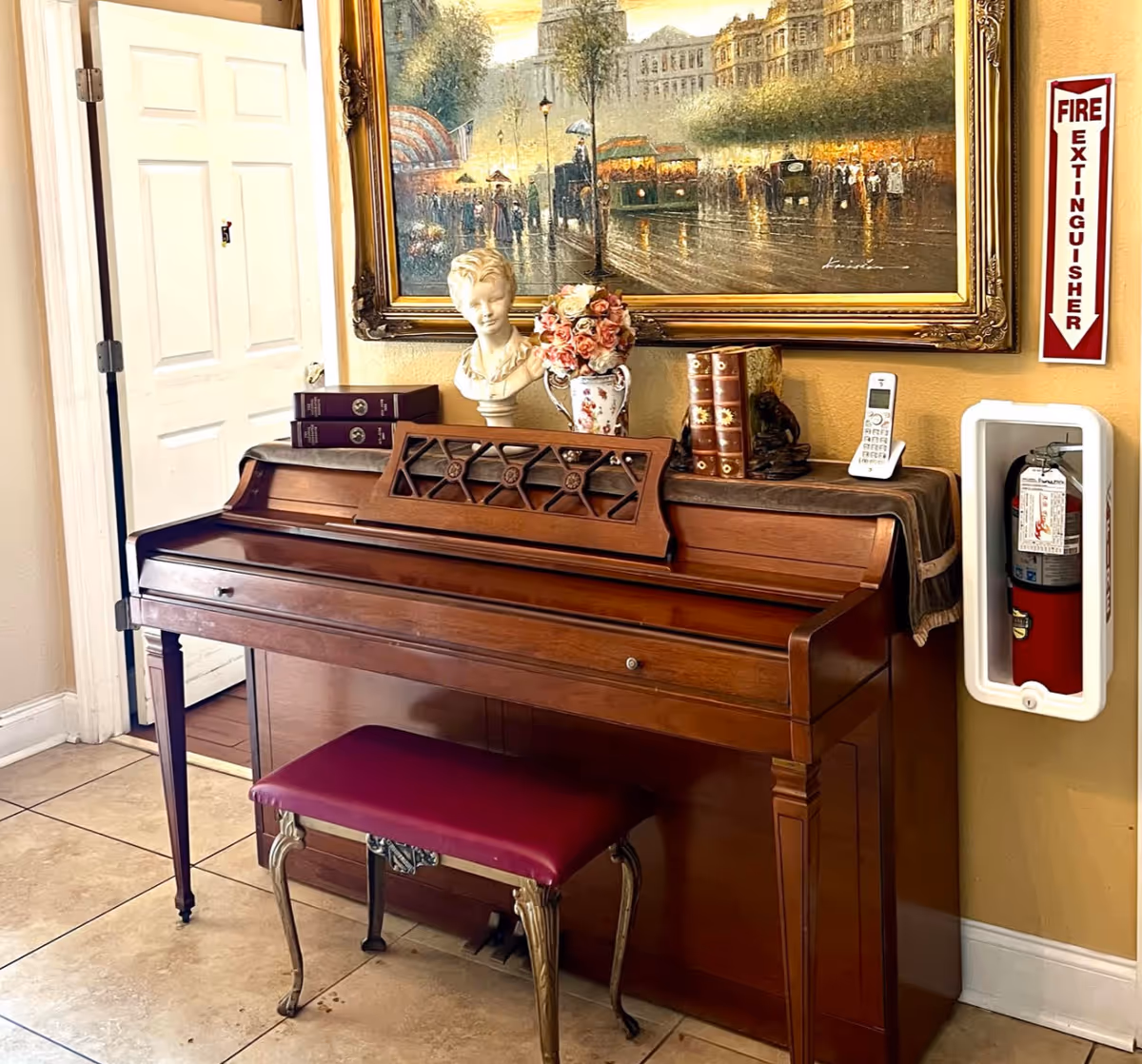 Wooden upright piano with a red cushioned bench, decorative bust, flowers and books on top, a framed painting above and a wall-mounted fire extinguisher nearby.