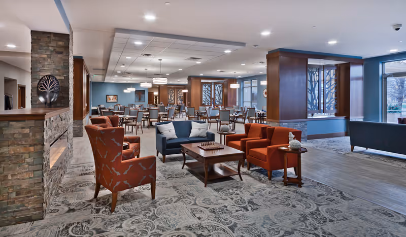 A spacious and well-lit common area in a senior living facility featuring a seating area with armchairs and a sofa around a coffee table with a chessboard. In the background, there are multiple dining tables and chairs arranged neatly. The room has a modern design with stone accents on the fireplace and decorative wooden panels on the walls, large windows letting in natural light, and pendant lights hanging from the ceiling.