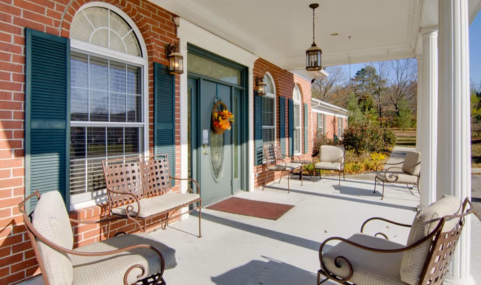 Covered porch area at Autumn Oaks with cushioned metal chairs and benches arranged around the entrance. The building has red brick walls, green shutters, and a green door decorated with a fall-themed wreath. Lantern-style light fixtures hang from the ceiling and are mounted on the walls. Trees and shrubs are visible in the background under a clear sky.
