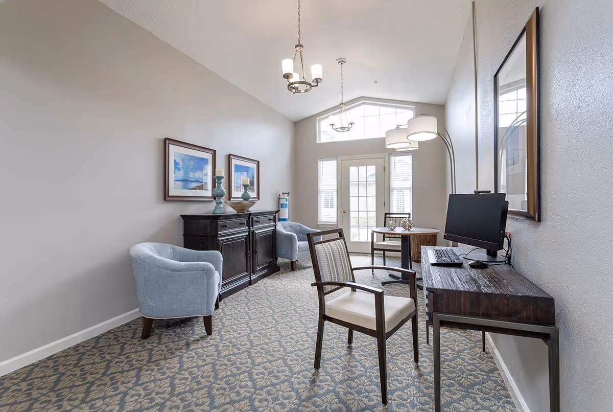 A bright and airy room with patterned carpet, light gray walls, and a high ceiling with two hanging light fixtures. The room features a dark wooden desk with a computer monitor, keyboard, and mouse, a wooden chair with a cushioned seat, two light blue upholstered armchairs, a dark wooden cabinet with decorative vases and framed artwork above it, and a round wooden table with two chairs near a glass door and windows letting in natural light.