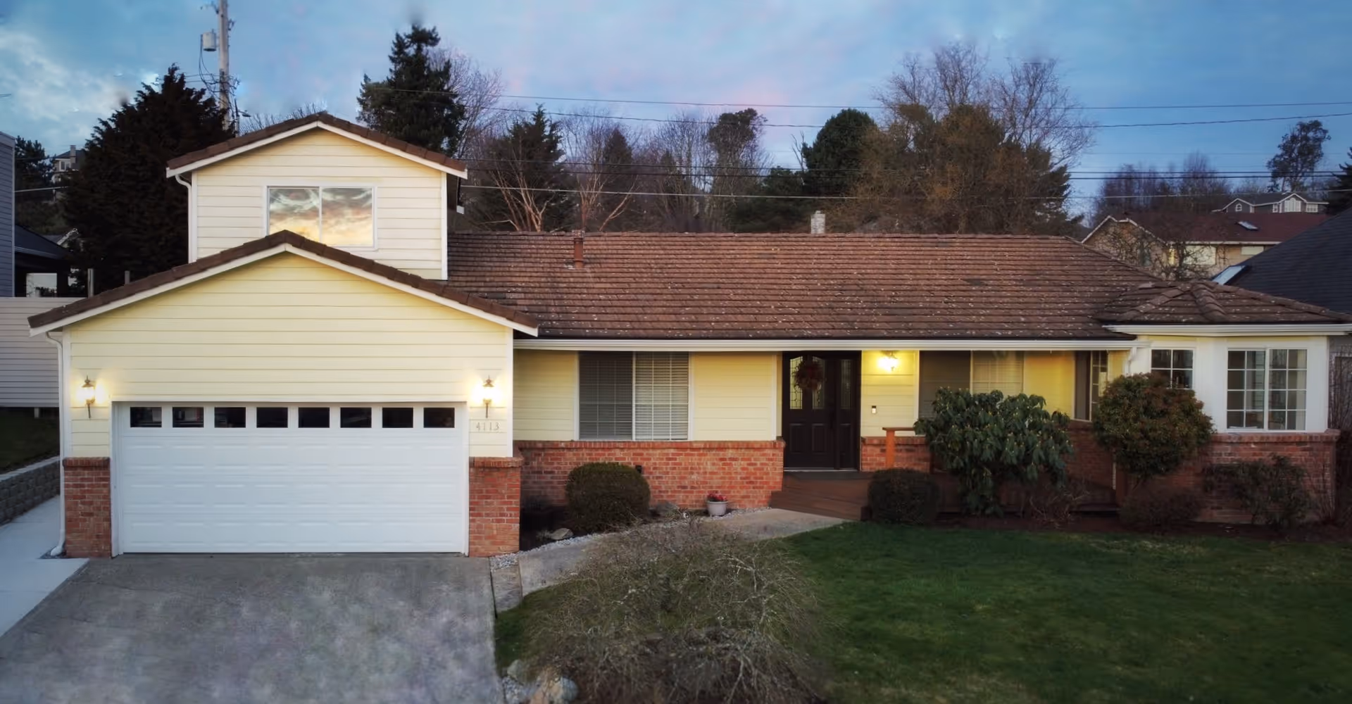 Exterior front view of a single-family home with a two-car garage, brick and yellow siding, a front porch with a door and windows, and a well-maintained lawn and shrubs in the foreground during dusk.