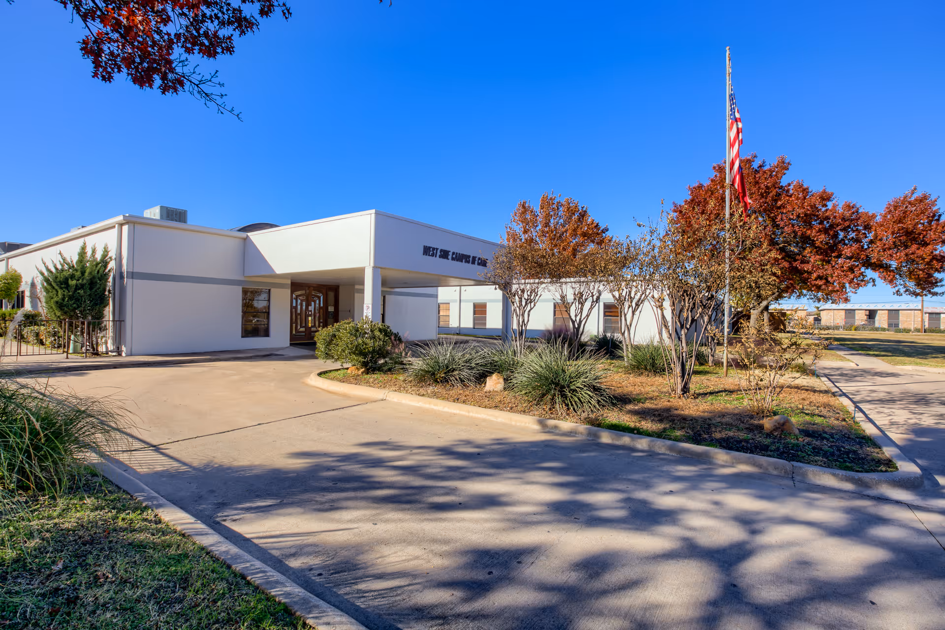 Exterior view of the West Side Campus of Care building on a clear sunny day, showing a driveway, landscaped bushes, trees with autumn foliage, and an American flag on a flagpole.