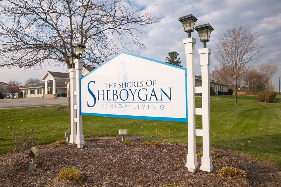 Outdoor view of a white and blue sign for The Shores of Sheboygan Senior Living, mounted on two white posts with lantern-style lights on top, surrounded by grass, trees, and a building in the background under a partly cloudy sky.
