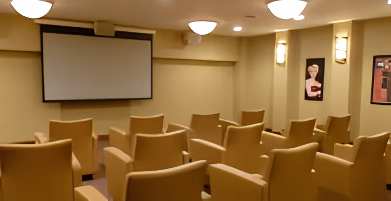 Small screening room with rows of beige armchairs facing a wall-mounted projection screen under soft ceiling lights.