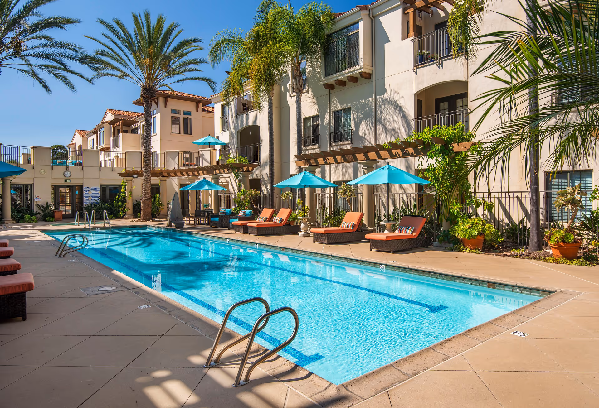Sunny outdoor swimming pool courtyard with lounge chairs, turquoise umbrellas, palm trees, and a multi-story residential building.
