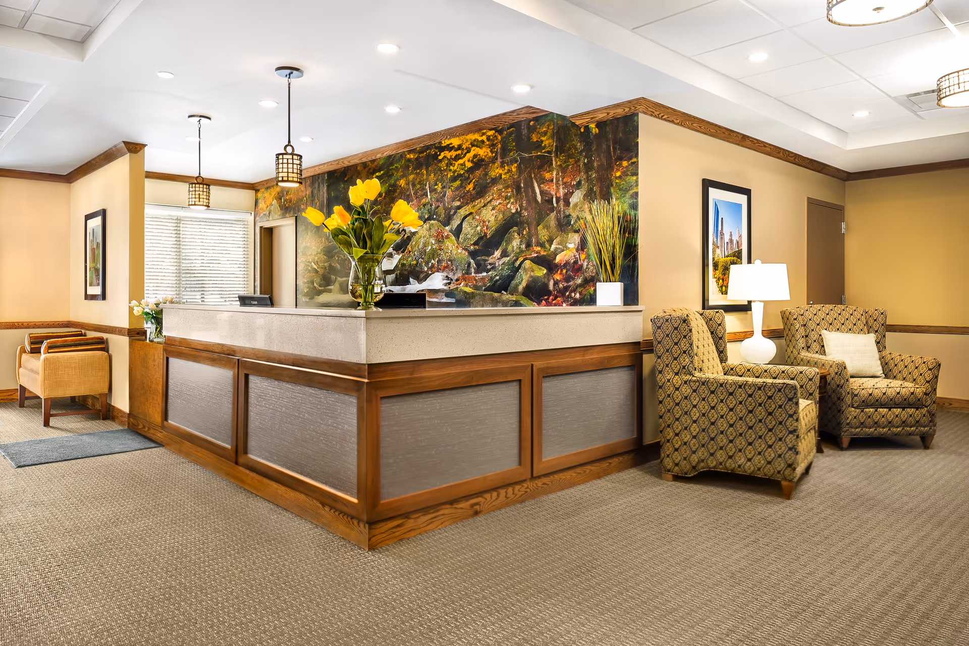 Reception area of a senior living facility with a wooden front desk decorated with a vase of yellow flowers. Behind the desk is a large mural depicting a forest scene with rocks and autumn foliage. To the right, there are two patterned armchairs with a white lamp on a small table between them. The walls are painted beige with wood trim, and there are framed pictures hanging on the walls. The floor is carpeted in a neutral tone.