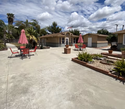 Outdoor patio area with concrete flooring, red tables and chairs with red umbrellas, surrounded by small plants and palm trees under a partly cloudy sky. There are single-story buildings in the background with an American flag flying on a pole.