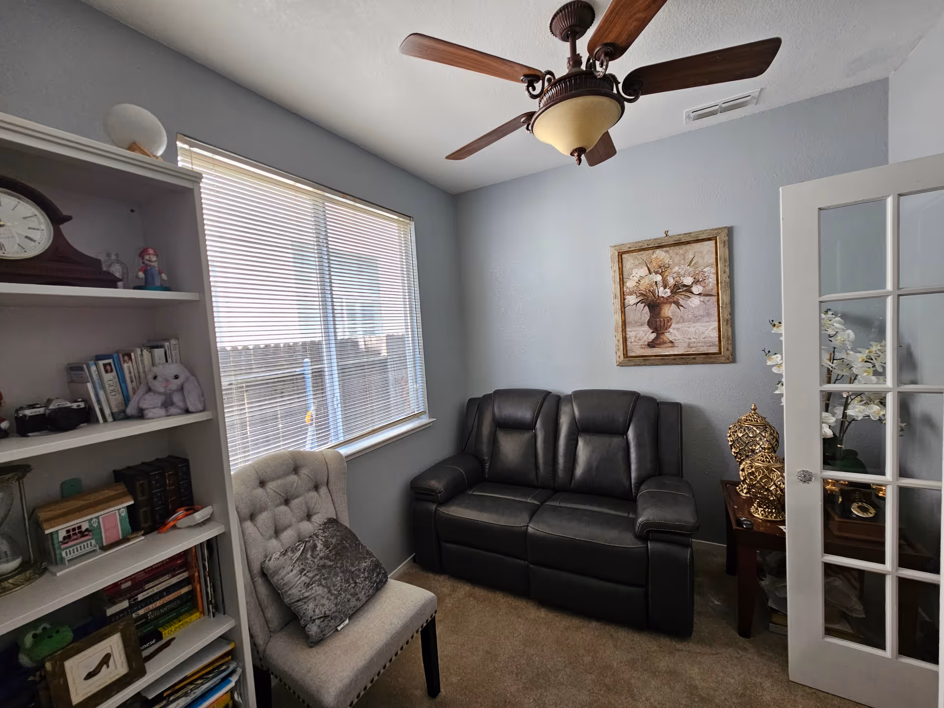 A small cozy room with a black leather loveseat, a beige tufted chair with a gray pillow, a wooden bookshelf filled with books and decorative items, a ceiling fan with wooden blades, a window with blinds, a framed floral painting on the wall, and a small wooden side table with a decorative golden object and white flowers.