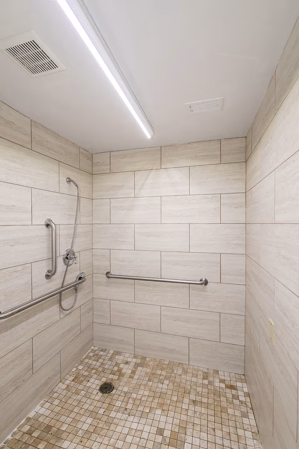 A tiled shower area with beige rectangular wall tiles and small square floor tiles in various shades of beige and brown. The shower features a handheld showerhead attached to the wall, two horizontal stainless steel grab bars for support, and a drain in the center of the floor. The ceiling has a long fluorescent light fixture and ventilation vents.
