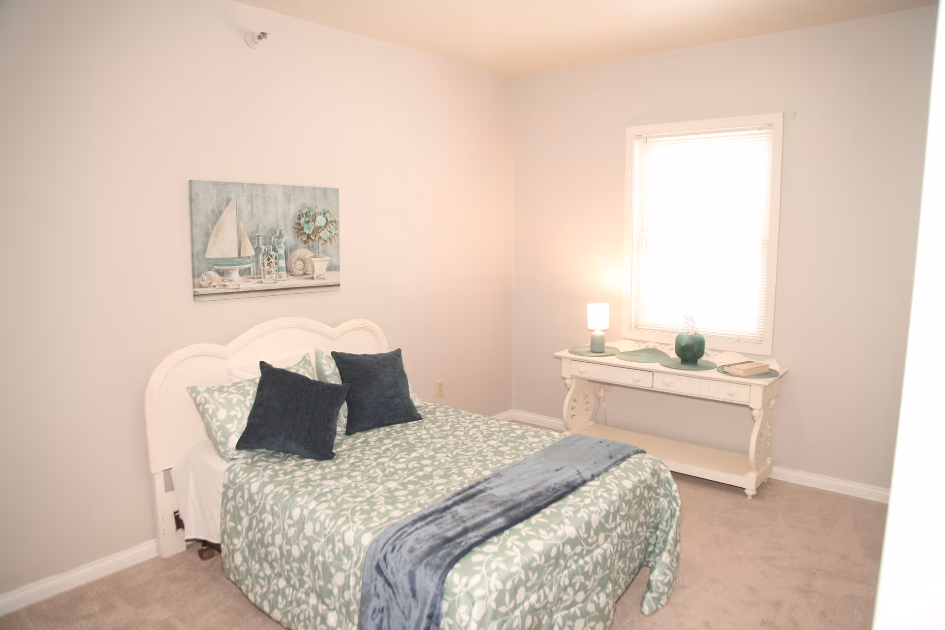 A small bedroom with a bed covered in a green and white floral bedspread, two dark blue pillows, and a blue throw blanket. There is a white headboard and a painting of a sailboat and seashells on the wall above the bed. A white table with a lamp, a green vase, and a book is positioned under a window with blinds.
