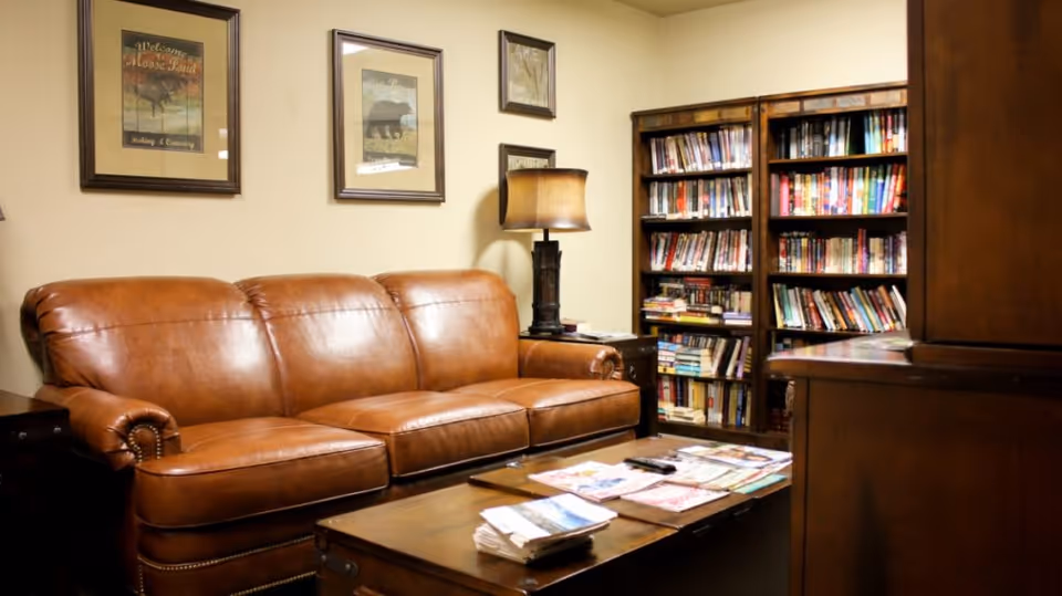 A cozy living room area with a brown leather sofa, a wooden coffee table with magazines on it, a table lamp on a side table, and bookshelves filled with books against the wall. Three framed pictures hang on the wall above the sofa.